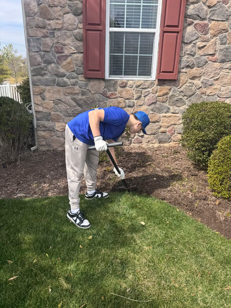 Person wearing blue shirt and cap digging soil in a garden bed beside a stone house wall with red shutters.
