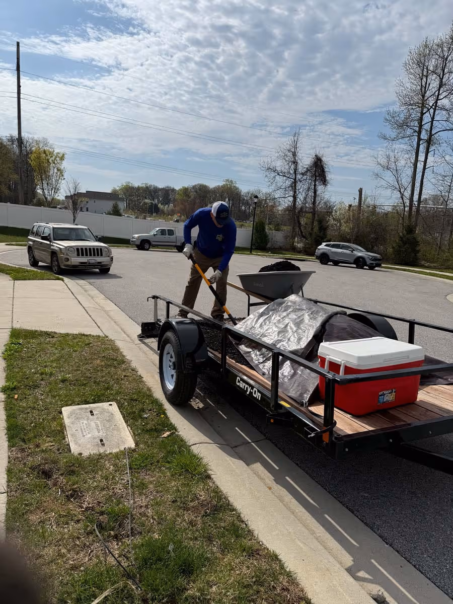 Person wearing blue shirt and cap shoveling soil from a trailer parked on the street.