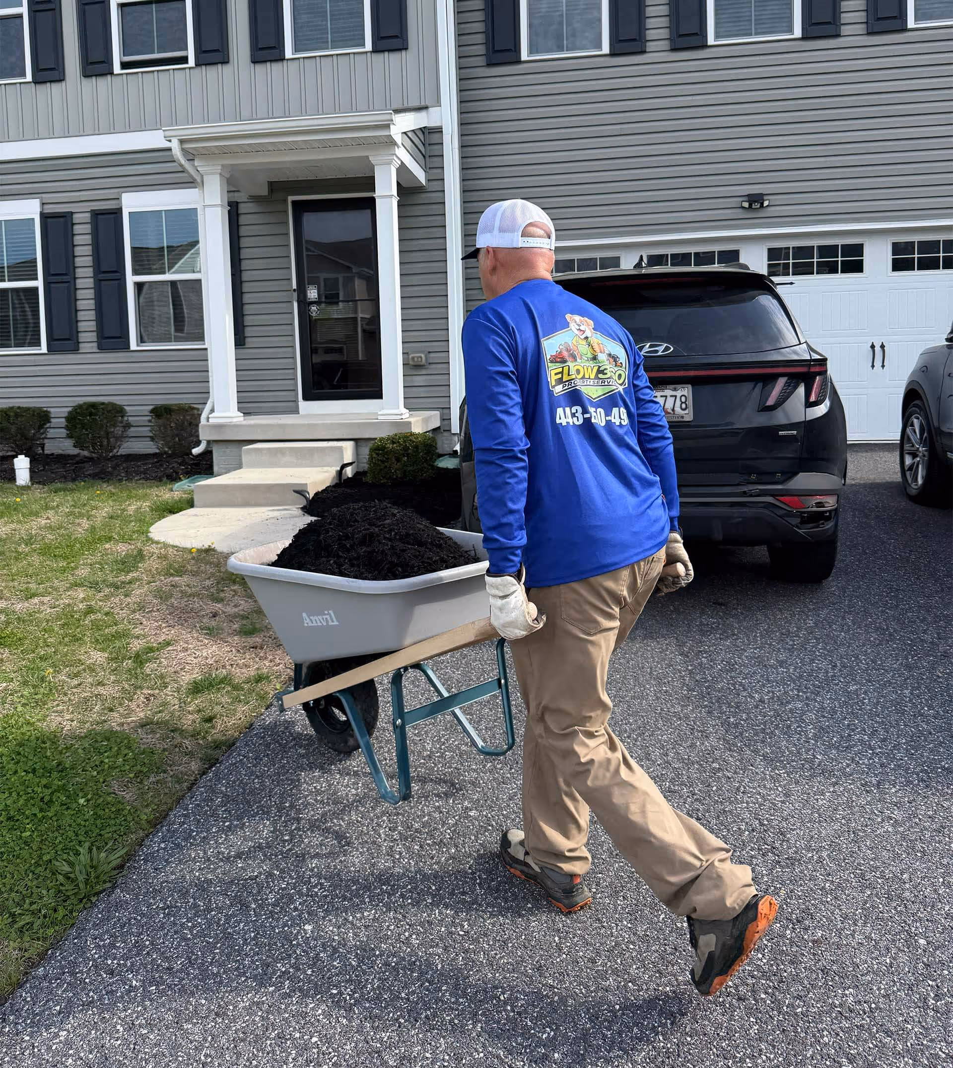 Man wearing a blue Flow Pro Services shirt pushing a wheelbarrow full of black mulch on a driveway in front of a gray house.