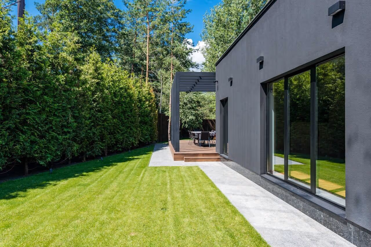 Modern house with dark exterior wall, large windows, concrete pathway, lawn, and patio with outdoor furniture under a pergola.