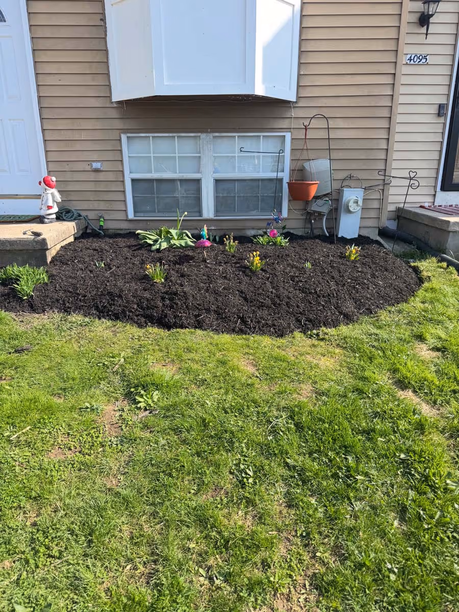 Front garden bed with dark mulch, several small plants, a pink flamingo decoration, and a hanging orange pot against a beige house wall.