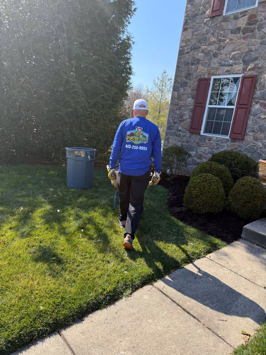 Person walking on grass near a stone house carrying garden tools, wearing a blue shirt with landscaping company logo and phone number.
