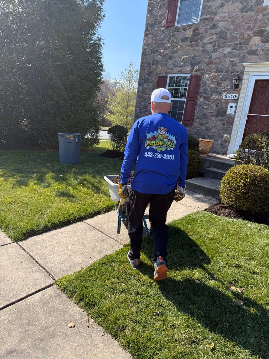 Person in a blue Flow360 shirt pushing a wheelbarrow on a sidewalk beside a stone house with a red door.