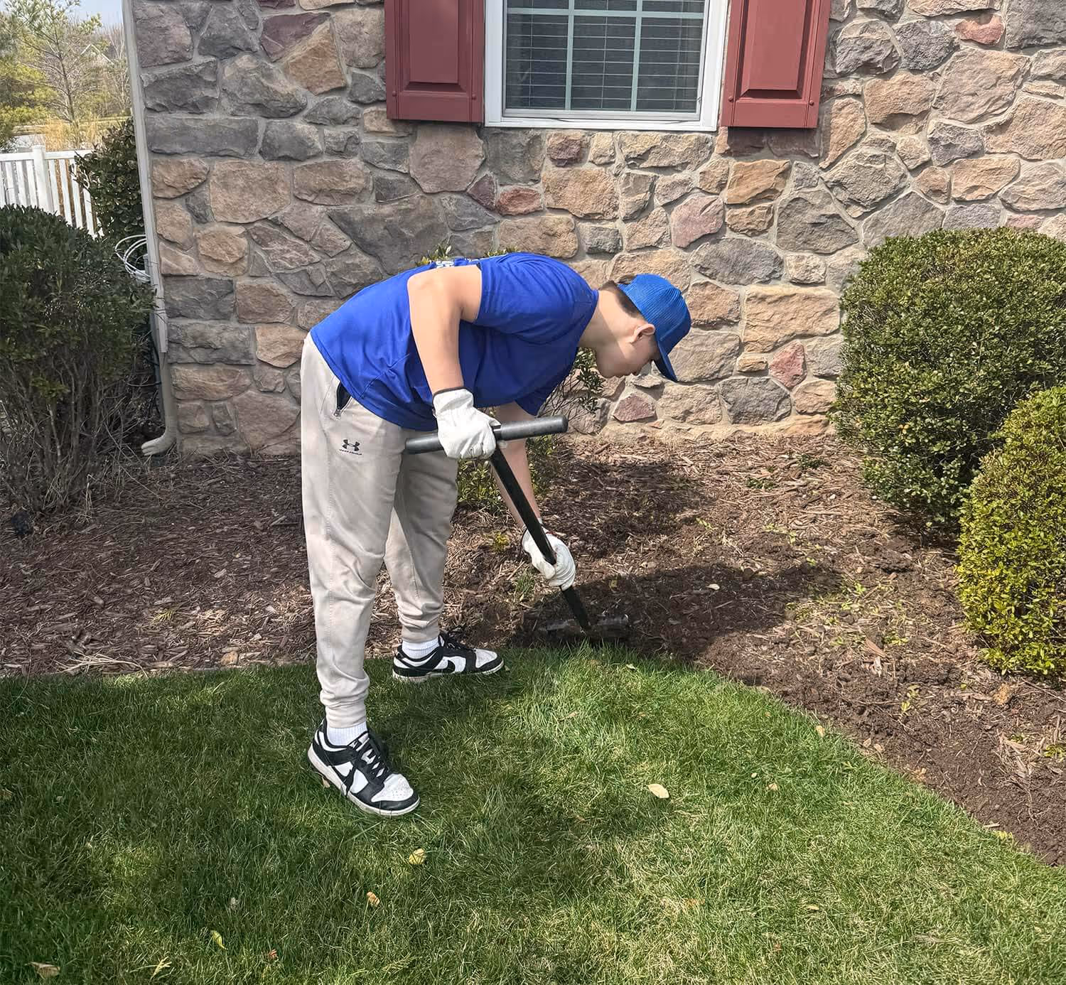 Person in blue shirt and cap using a gardening tool to work in a garden bed next to a stone wall house.