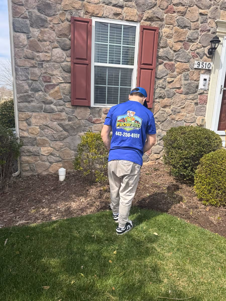 Person wearing a blue FLOW360 T-shirt working on landscaping near a stone house with red shutters and green bushes.